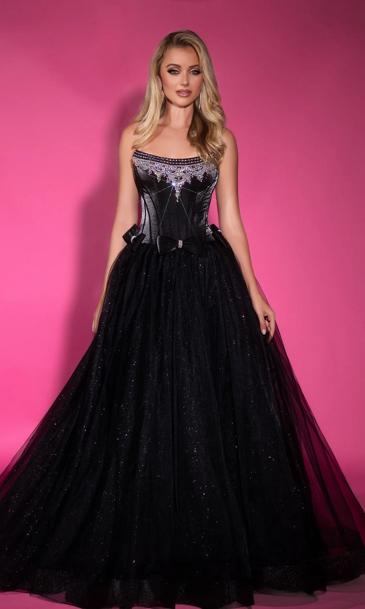 A woman stands in front of a pink background wearing the Portia and Scarlett PS25336 long formal dress in black, featuring a strapless corset bodice and a full glitter tulle skirt.